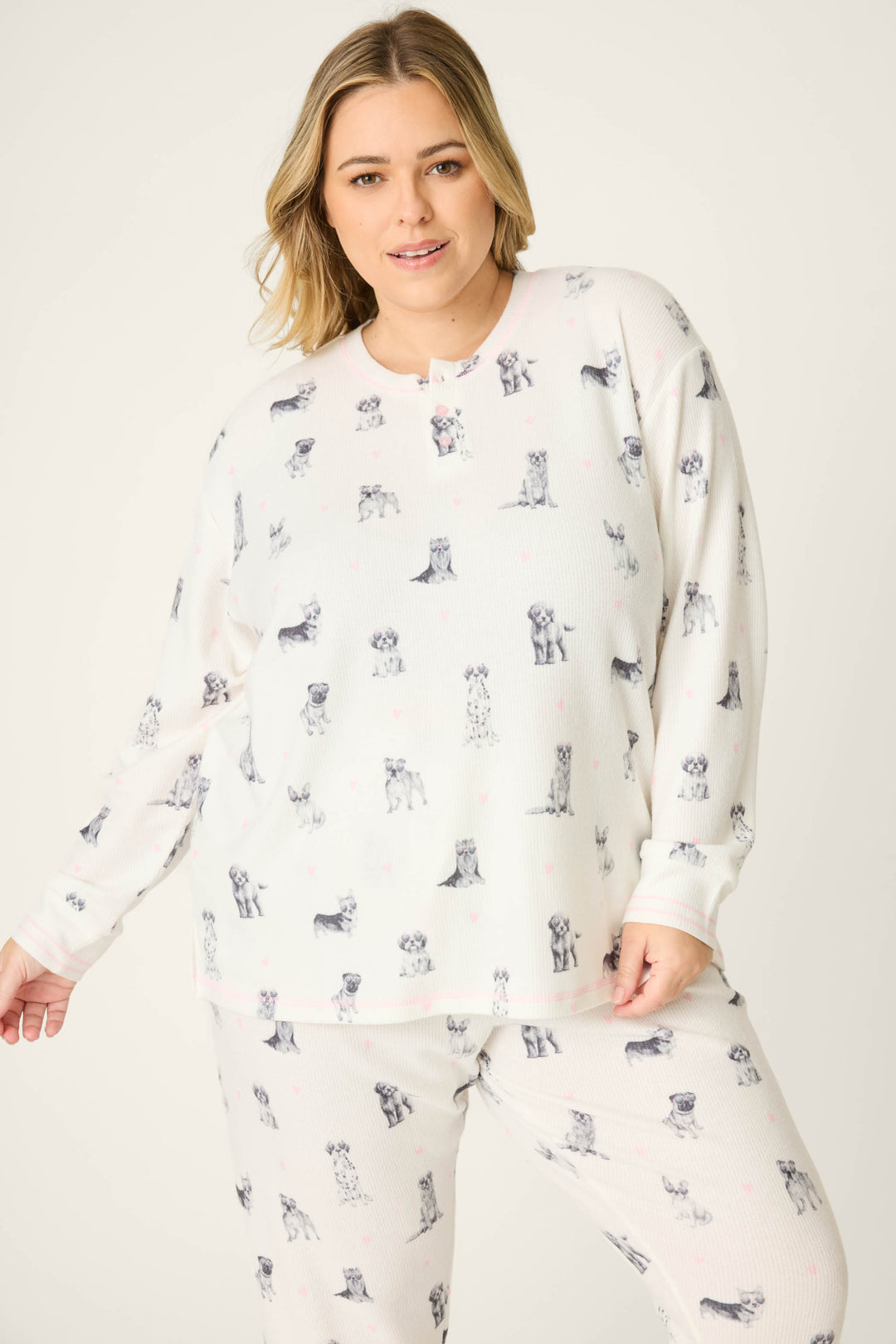 A woman models the P.J. Salvage "My Dog Is My Valentine" Plus Size PJ Set, featuring a white dog-print design, as she stands against a plain background and smiles softly at the camera.