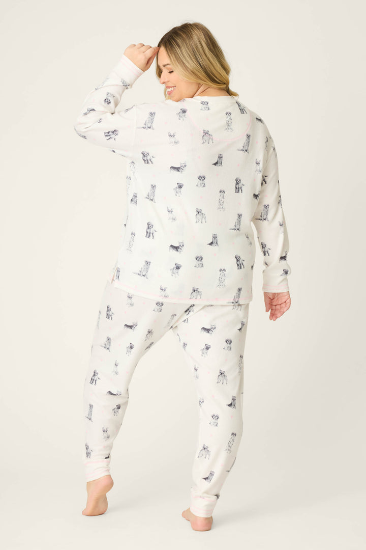 A woman stands barefoot facing away, wearing the P.J. Salvage "My Dog Is My Valentine" Plus Size PJ Set with a dog print, one hand raised to her head, posing against a plain light background.