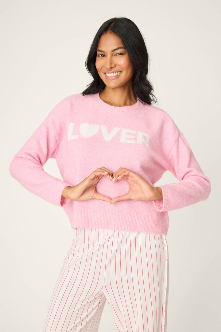 A person smiles, wearing the P.J. Salvage Feeling Loved "Lover" Snuggle Sweater in light pink, featuring "LOVER" with a heart for the O, and holds their hands in front, forming a heart shape with their fingers.