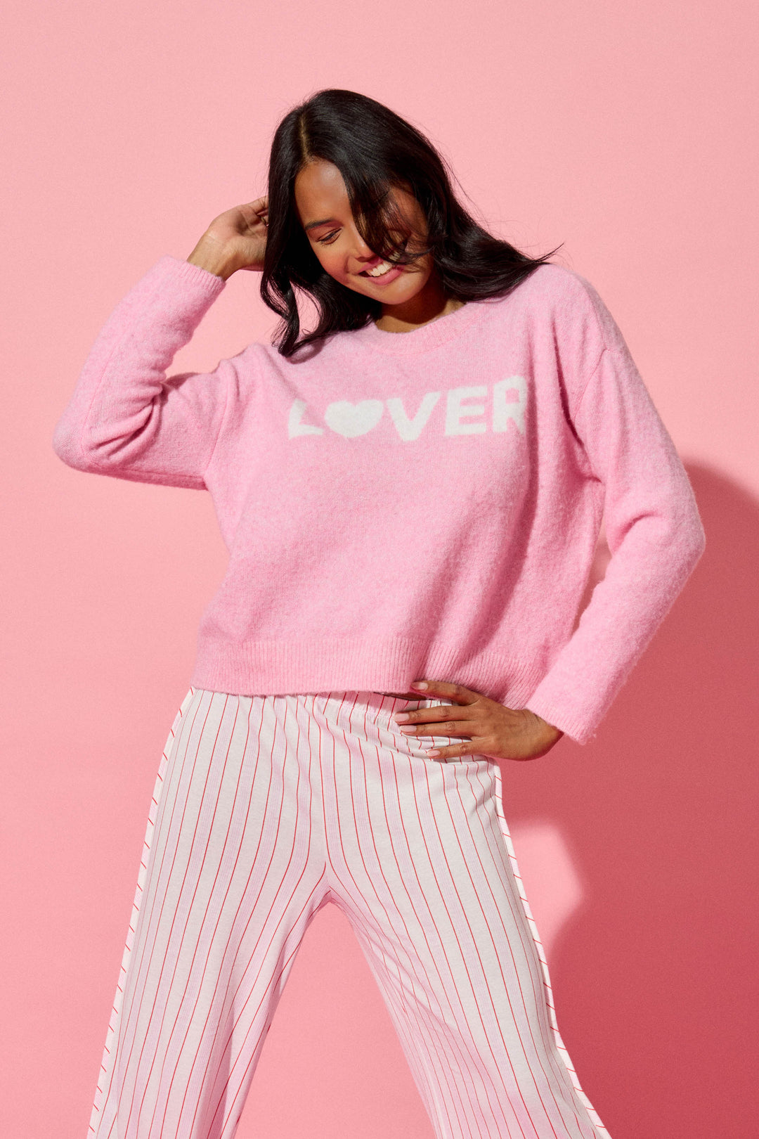 A person smiles and tilts their head against a pink background, wearing the P.J. Salvage Feeling Loved "Lover" Snuggle Sweater, paired with white pants featuring thin red vertical stripes.