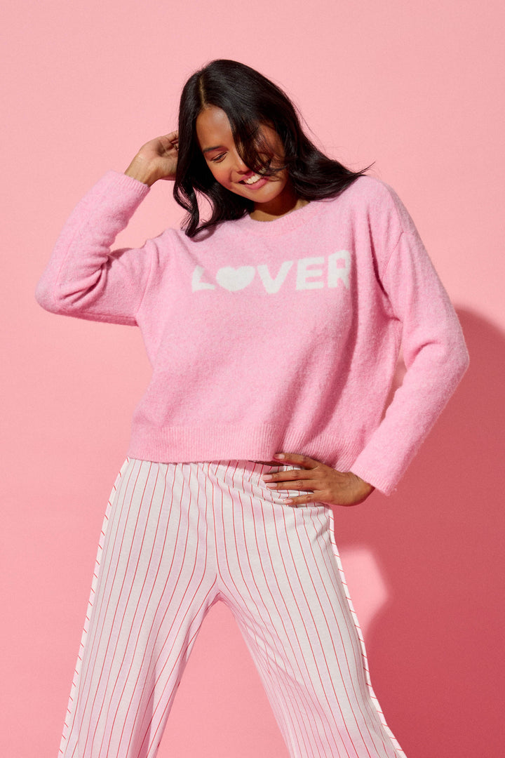 A person smiles and tilts their head against a pink background, wearing the P.J. Salvage Feeling Loved "Lover" Snuggle Sweater, paired with white pants featuring thin red vertical stripes.