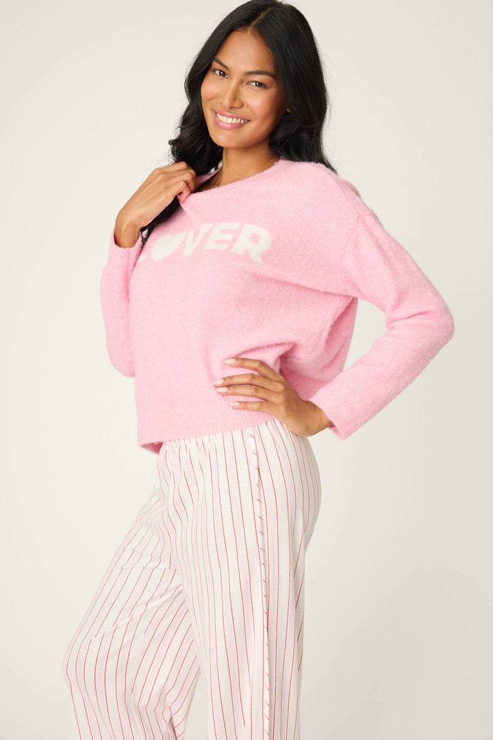 A person smiles, wearing the P.J. Salvage Feeling Loved "Lover" Snuggle Sweater in light pink with white lettering, paired with white pants with red stripes, posing with one hand on their hip and the other holding the sweater collar.