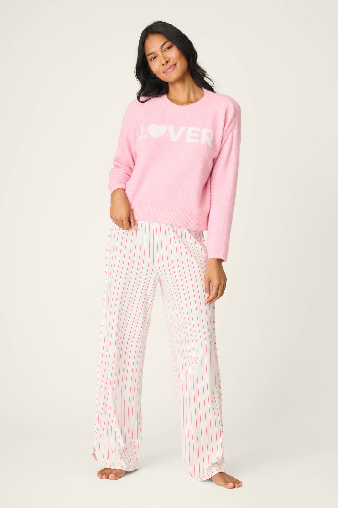 A woman stands barefoot in the P.J. Salvage Feeling Loved "Lover" Snuggle Sweater, a pale pink top with white lettering, paired with loose white pants featuring thin red stripes, posing against a plain light background.