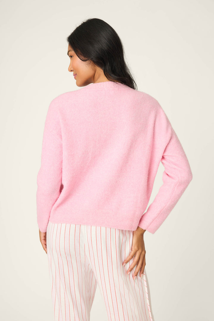 A person with long dark hair stands with their back to the camera, wearing the P.J. Salvage Feeling Loved "Lover" Snuggle Sweater in light pink and white pants with red stripes against a plain light background.