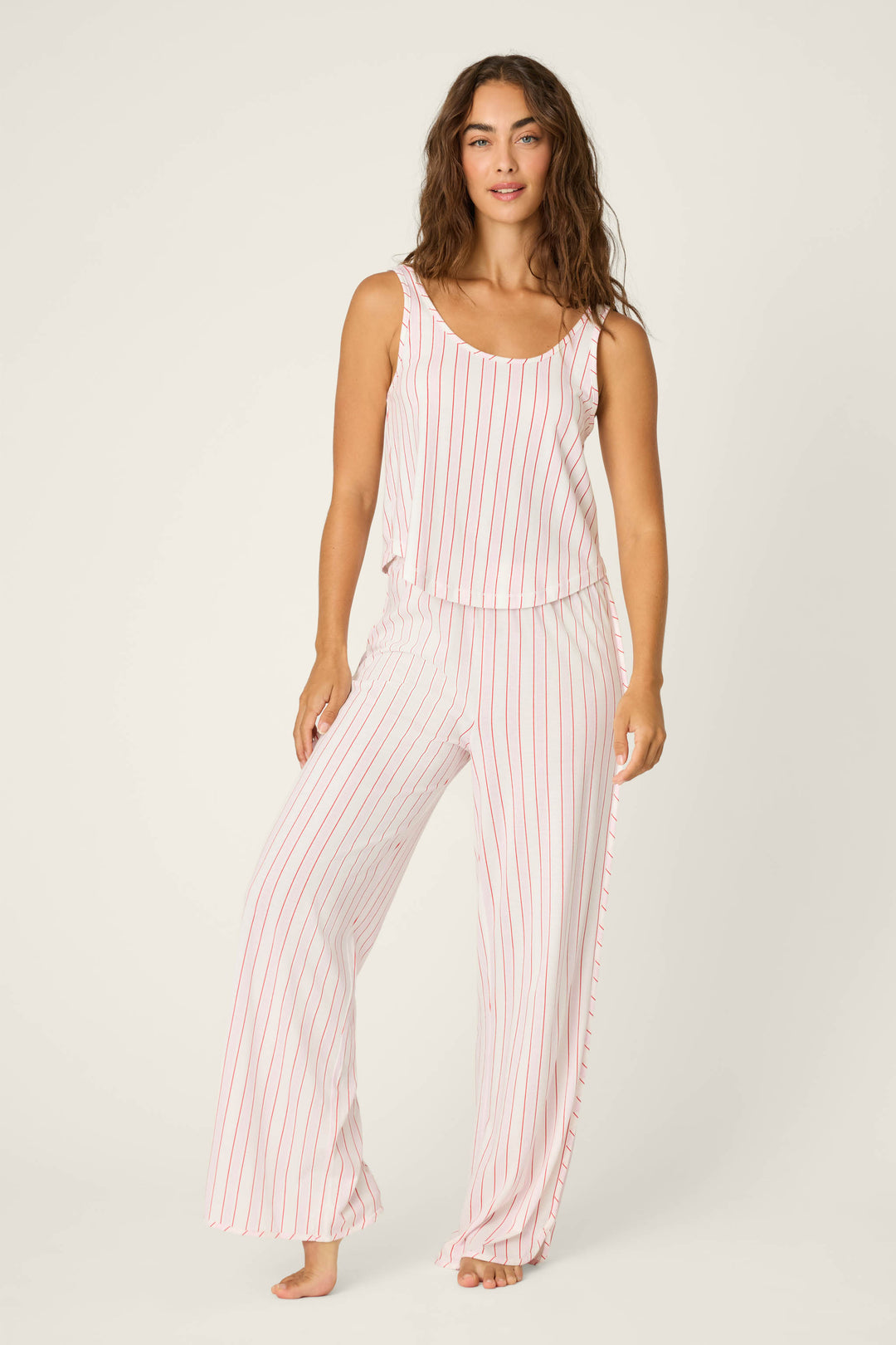 A woman stands barefoot against a plain background, wearing P.J. Salvage’s Feeling Loved Striped Open Leg Pants—comfy pajama pants featuring vertical pink stripes on soft white fabric.