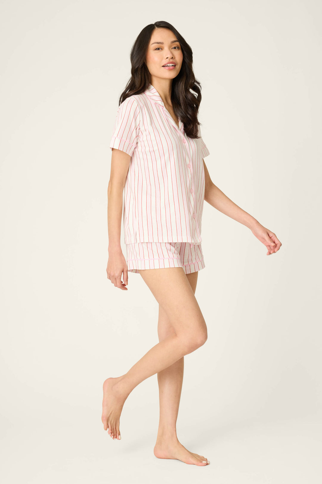 A woman with long, dark hair stands barefoot against a plain background, modeling the Feeling Love Striped 3-Piece PJ Set by P.J. Salvage—light-colored pajamas with vertical pink stripes—one leg bent, arms relaxed at her sides.