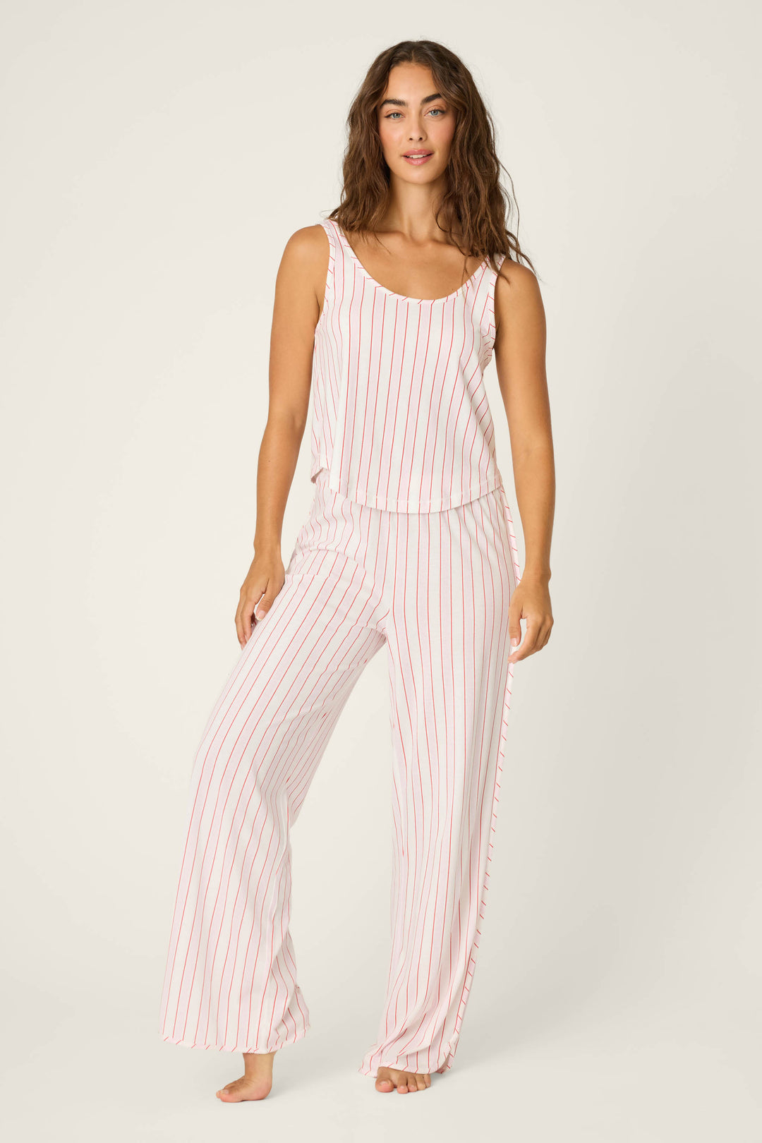 A woman stands barefoot against a plain light background wearing the P.J. Salvage Feeling Loved Striped PJ Set—a sleeveless top and wide-leg pants made of white fabric with vertical red stripes.