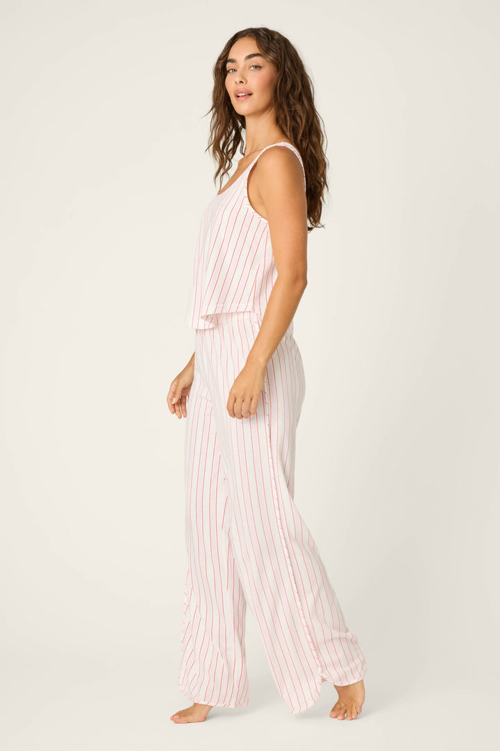 A person with long, wavy brown hair stands barefoot against a plain light background, wearing the P.J. Salvage Feeling Loved Striped PJ Set—a sleeveless, loose white pajama with vertical red stripes.