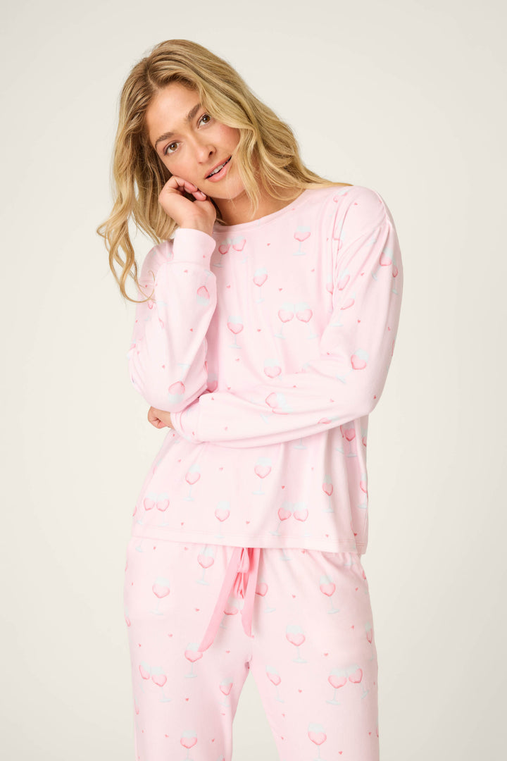 A person stands against a plain background, wearing the P.J. Salvage Happy Hour Wine Glass Print Long Sleeve Top and matching pants. Their head is tilted, with one hand resting on their cheek.