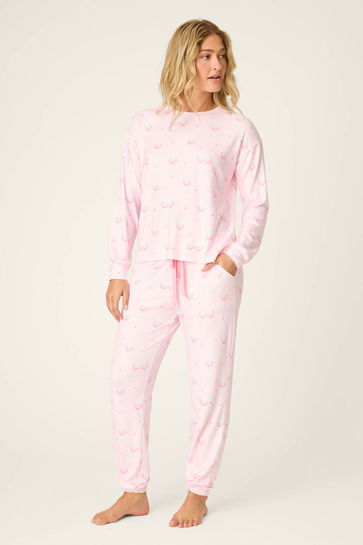 A person stands barefoot in a relaxed-fit P.J. Salvage Happy Hour Wine Glass Print Long Sleeve Top and matching pants, featuring a playful wine glass pattern. The background is plain light beige.