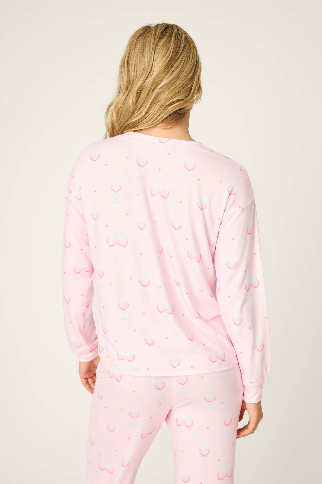 A person with long blonde hair stands with their back to the camera, wearing the P.J. Salvage Happy Hour Wine Glass Print Long Sleeve Top in a light pink shade. The background is plain and neutral.