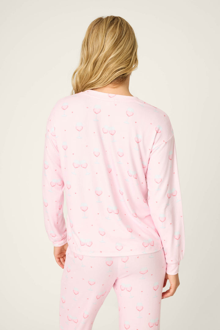 A person with long blonde hair stands with their back to the camera, wearing the P.J. Salvage Happy Hour Wine Glass Print Long Sleeve Top in a light pink shade. The background is plain and neutral.