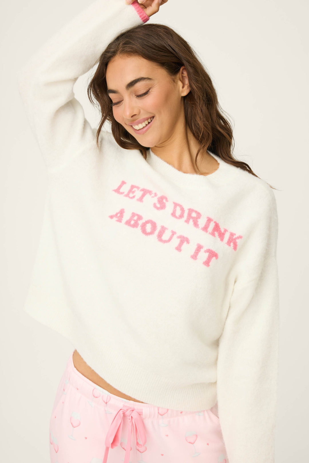 A woman smiles with one arm raised, wearing the P.J. Salvage Happy Hour "Lets Drink About it" Snuggle Sweater and light pink cherry-print pajama pants.
