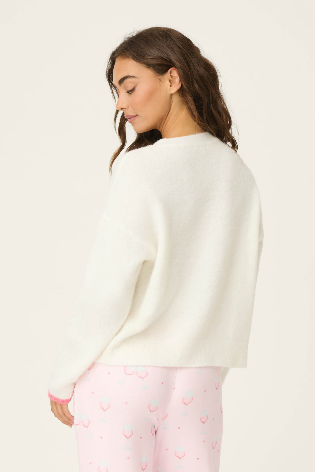 A woman faces away, glancing over her shoulder in a white P.J. Salvage Happy Hour "Let's Drink About it" Snuggle Sweater and light pink cherry-print pajama pants, set against a plain, light background.