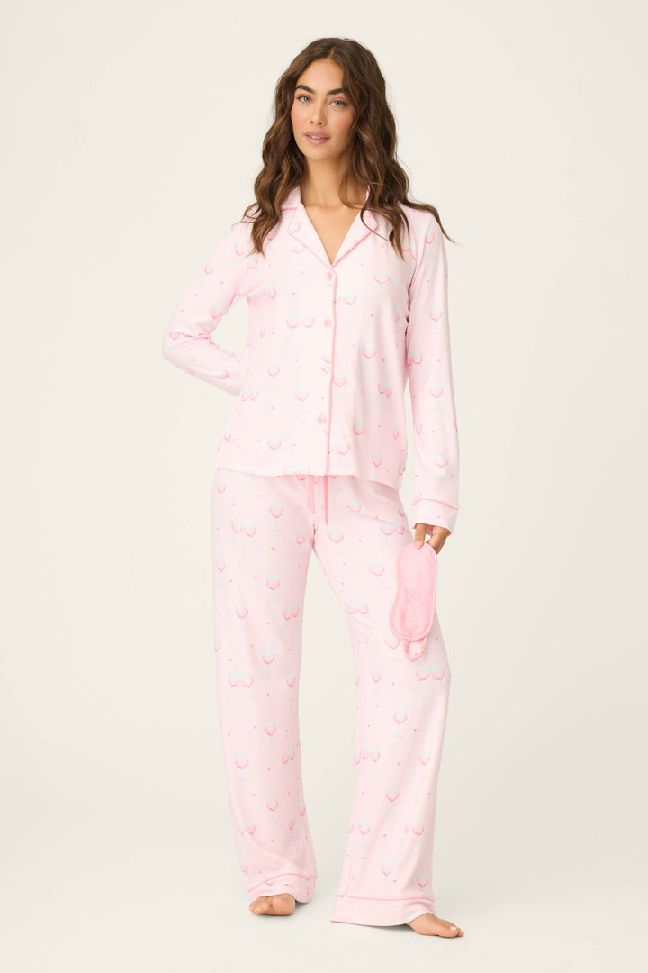A woman stands barefoot against a light background wearing the P.J. Salvage Happy Hour Wine Glass Print 3-Piece PJ Set in pink, holding the matching sleep mask and looking at the camera with a neutral expression.