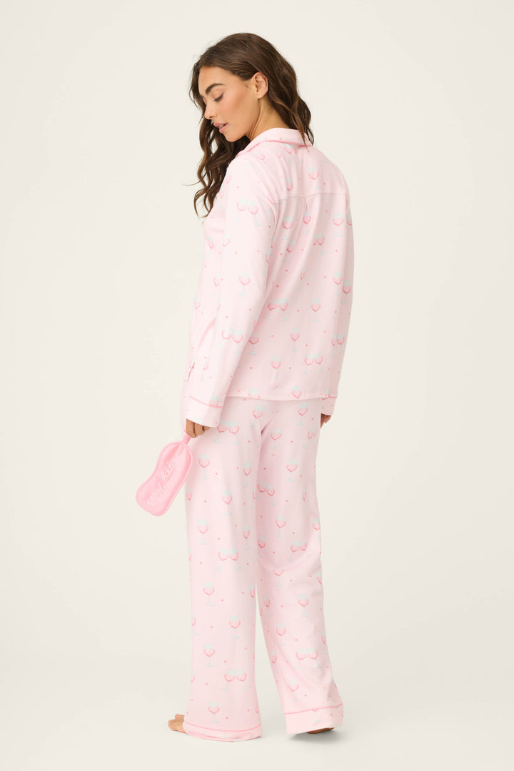 A person stands barefoot, facing slightly away, wearing the P.J. Salvage Happy Hour Wine Glass Print 3-Piece PJ Set and holds matching slippers in one hand against a plain, light background.