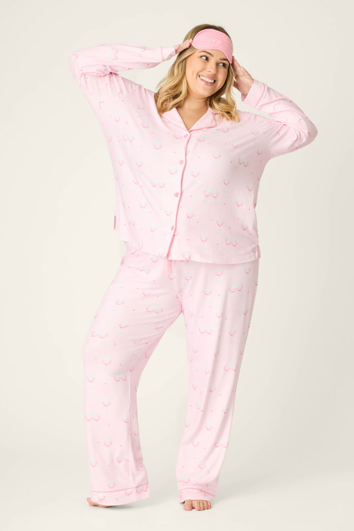 A person smiles while standing barefoot in the P.J. Salvage Happy Hour Wine Glass Print 3-Piece Plus Size PJ Set, featuring a playful pattern and matching sleep mask, against a plain light background.