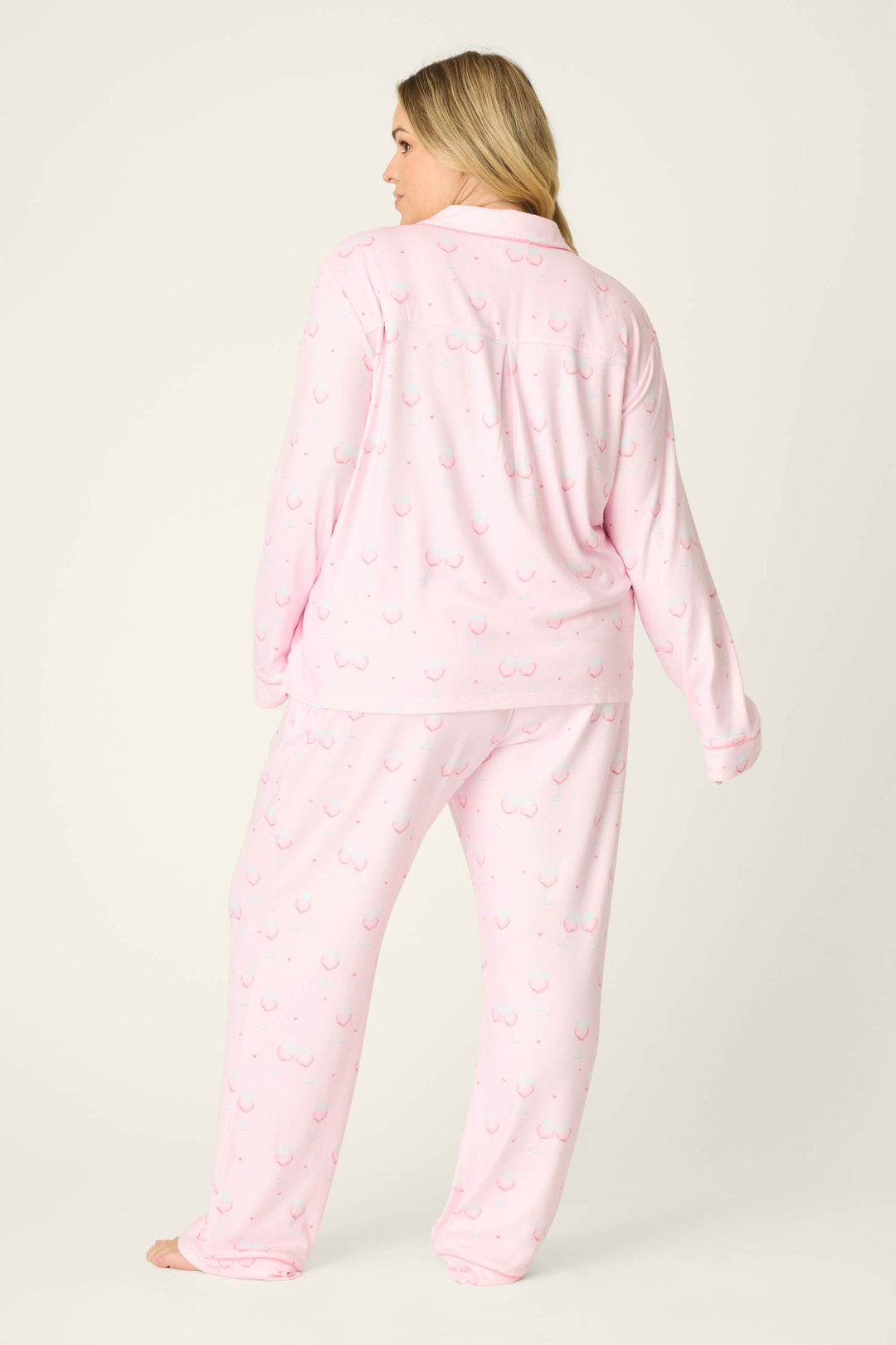 A person with long blonde hair stands barefoot, facing away, wearing the P.J. Salvage Happy Hour Wine Glass Print 3-Piece Plus Size PJ Set in light pink on a plain background.