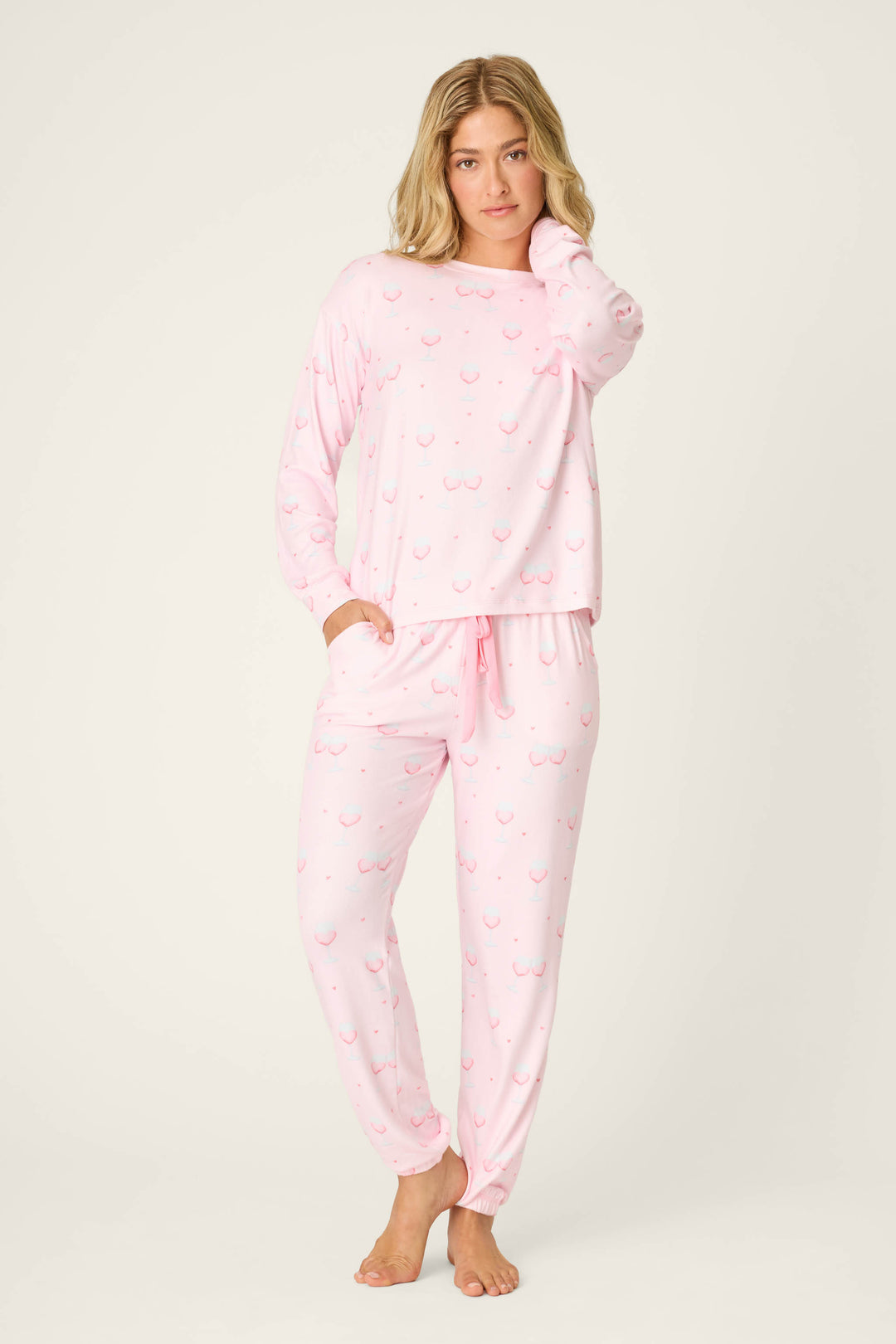A person stands barefoot on a neutral background, wearing the P.J. Salvage Happy Hour Wine Glass Print PJ Set—light pink long-sleeve pajamas with a small wine glass pattern, one hand on their face and the other in their pocket.
