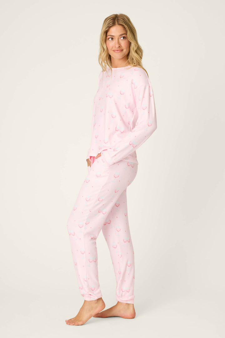 A person models the P.J. Salvage Happy Hour Wine Glass Print PJ Set, featuring a long-sleeved top and pants in light pink with a subtle red print, standing barefoot on a light background and facing slightly to the side.