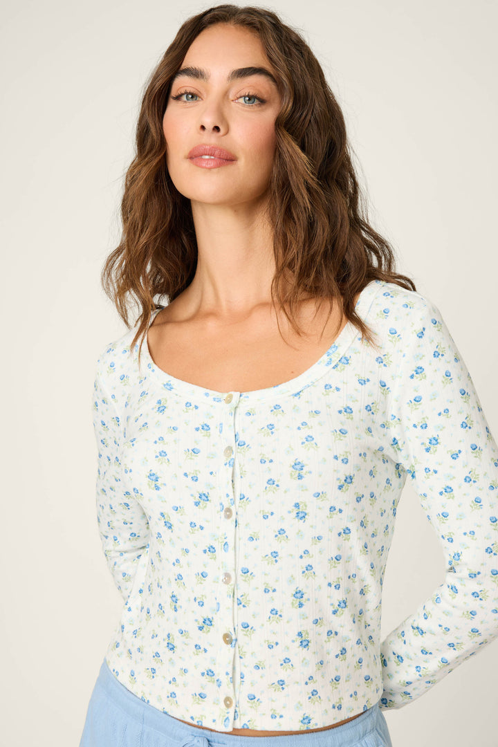 A woman with wavy brown hair models the P.J. Salvage More Amour Ditsy Floral Long Sleeve Pointelle Top, featuring a white base with a small blue floral pattern, against a plain, light background. She pairs it with light blue pants.