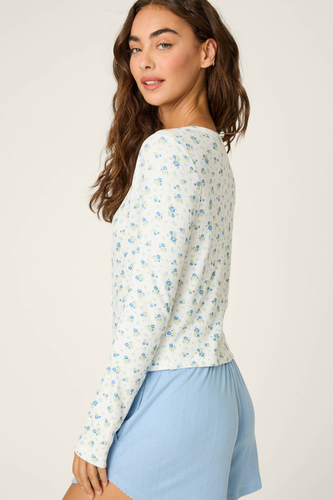 A woman with long brown hair stands sideways, wearing the P.J. Salvage More Amour Ditsy Floral Long Sleeve Pointelle Top and light blue shorts, glancing over her shoulder against a plain background.