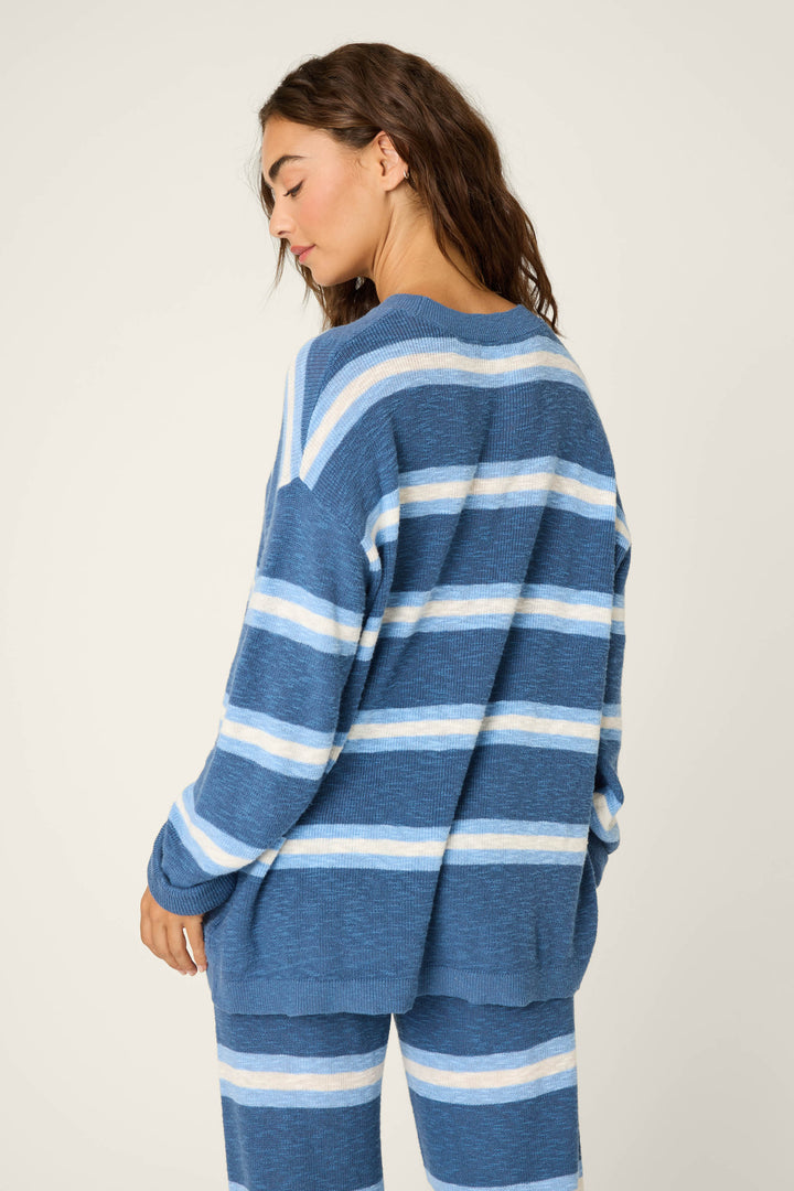 A woman with long brown hair faces away, wearing the P.J. Salvage More Amour Striped Long Sleeve Top—a loose blue knit sweater with horizontal white and light blue stripes—against a plain off-white background.