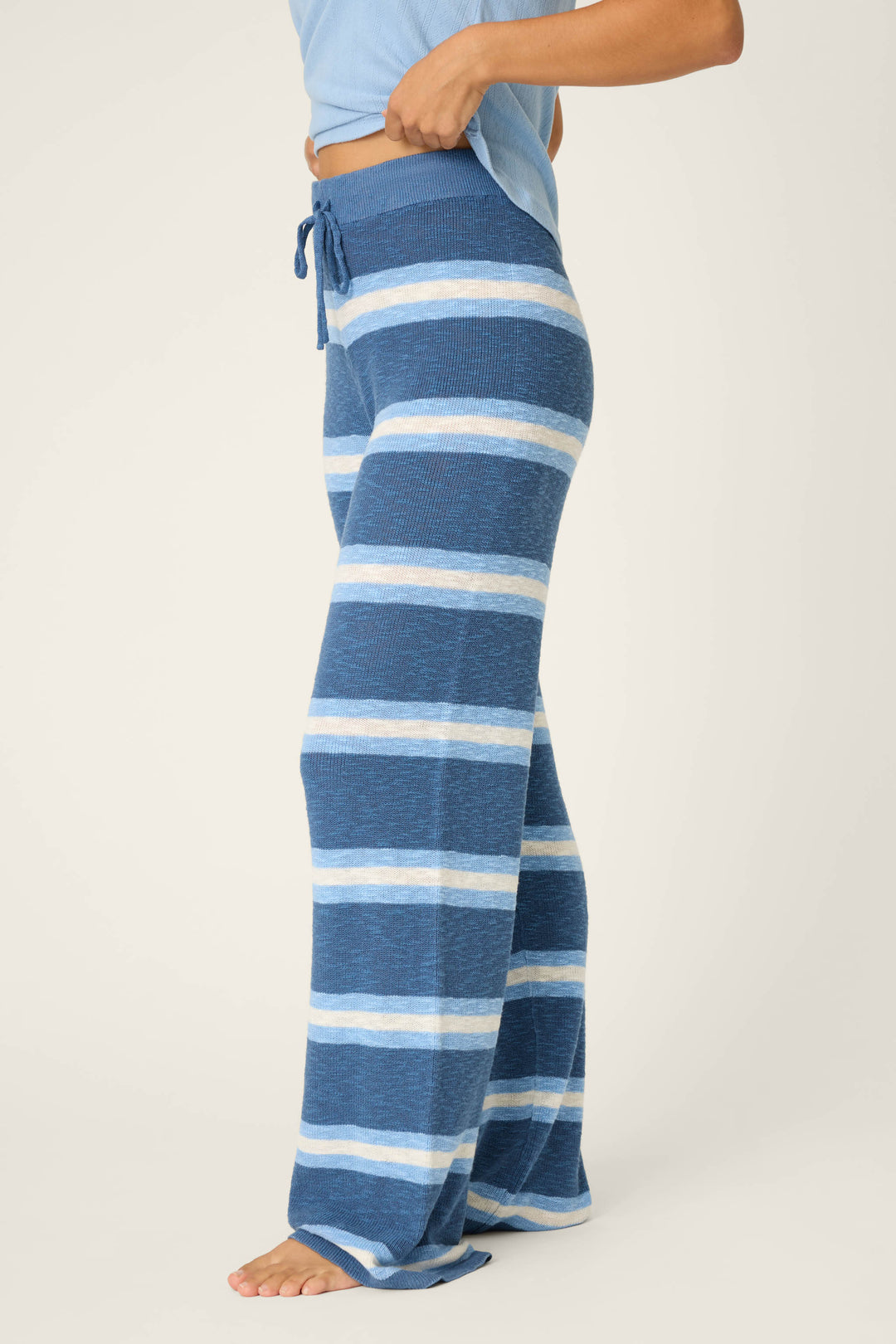 A person stands barefoot in P.J. Salvage's More Amour Striped Open Leg Pants, featuring blue and white stripes and a drawstring waist, shown from the side against a plain background.