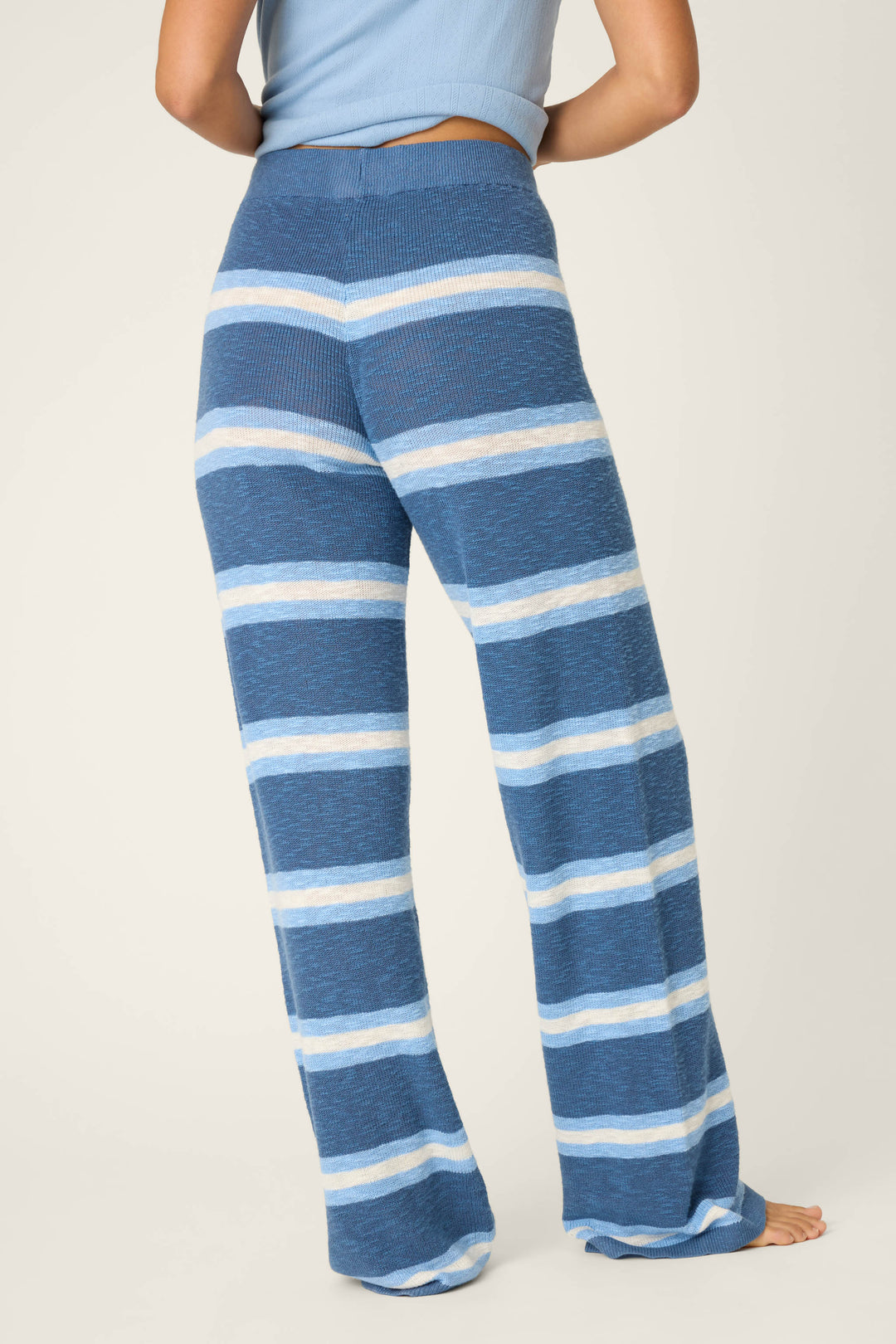 A person stands barefoot on a light background, wearing P.J. Salvage's More Amour Striped Open Leg Pants in blue and white stripes with a blue top. The full length of the pants is visible from behind.