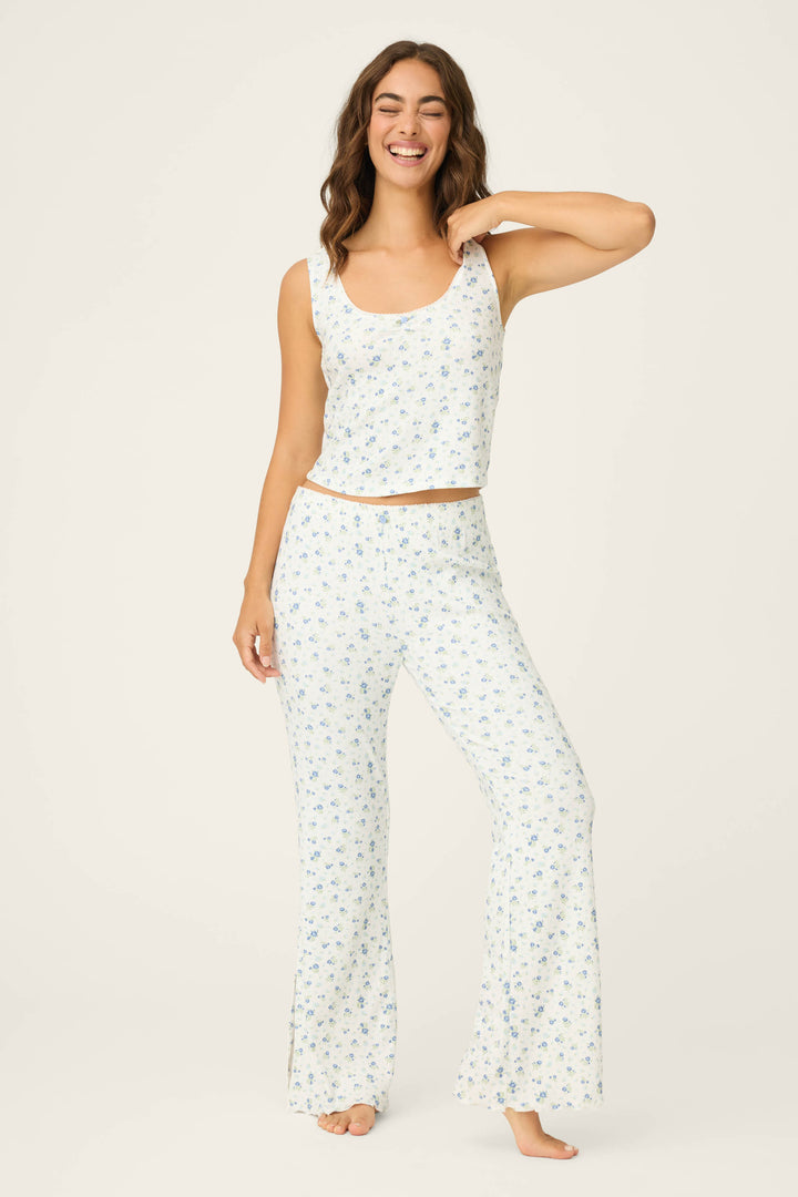 A woman stands barefoot on a light background, smiling in the P.J. Salvage More Amour Ditsy Floral Pointelle PJ Set—a matching white pajama set with a blue floral pattern, featuring a sleeveless top and long pants. One hand is raised near her neck.