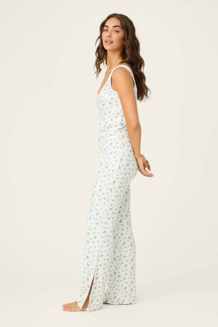 A woman with long wavy hair and a neutral expression stands in profile against a plain light background, wearing the P.J. Salvage More Amour Ditsy Floral Pointelle PJ Set—a sleeveless, long white set with a blue floral pattern and hem slit.