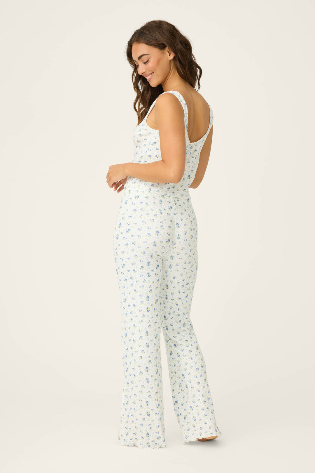 A woman smiles over her shoulder, wearing the P.J. Salvage More Amour Ditsy Floral Pointelle PJ Set—a white sleeveless set with a blue floral pattern—against a plain, light background.
