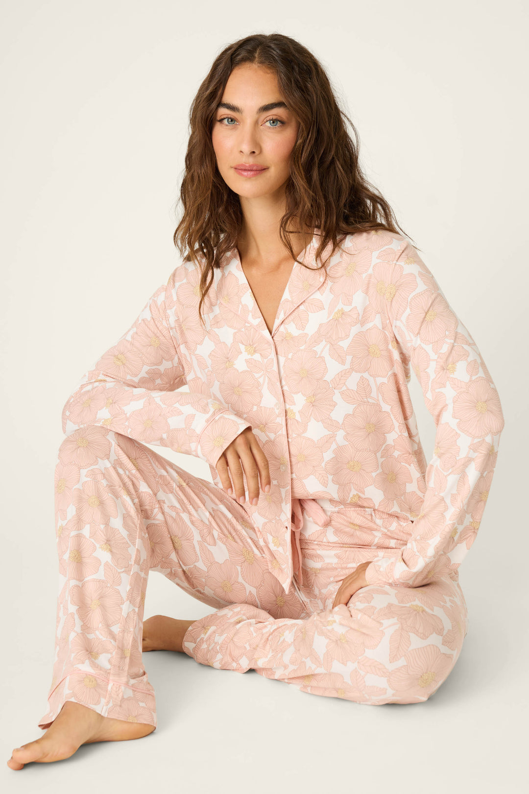 A woman with wavy brown hair sits on the floor with one knee up against a light background, wearing the Poppy Dreams Floral PJ Set by P.J. Salvage.