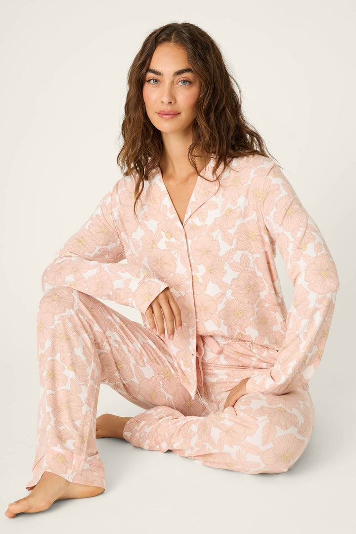 A woman with wavy brown hair sits on the floor with one knee up against a light background, wearing the Poppy Dreams Floral PJ Set by P.J. Salvage.