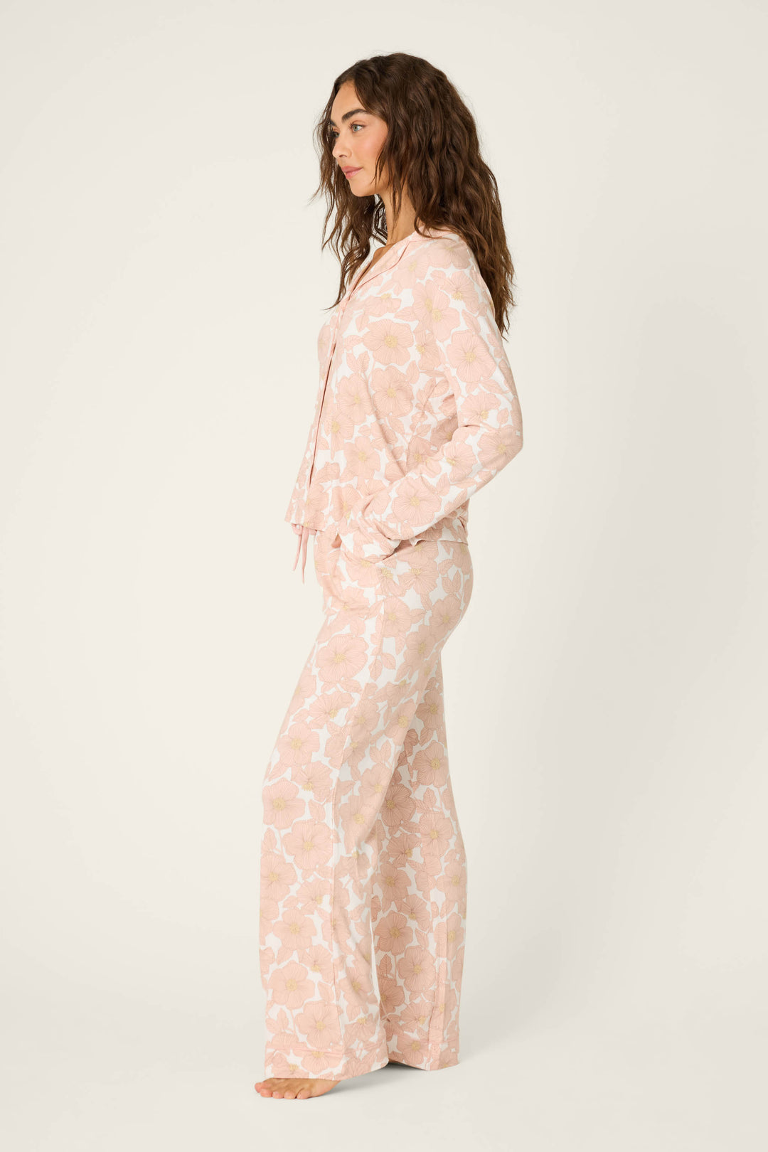 A woman stands barefoot in profile against a plain light background, wearing the P.J. Salvage Poppy Dreams Floral PJ Set—a matching long-sleeve top and pants featuring a light pink floral pattern.