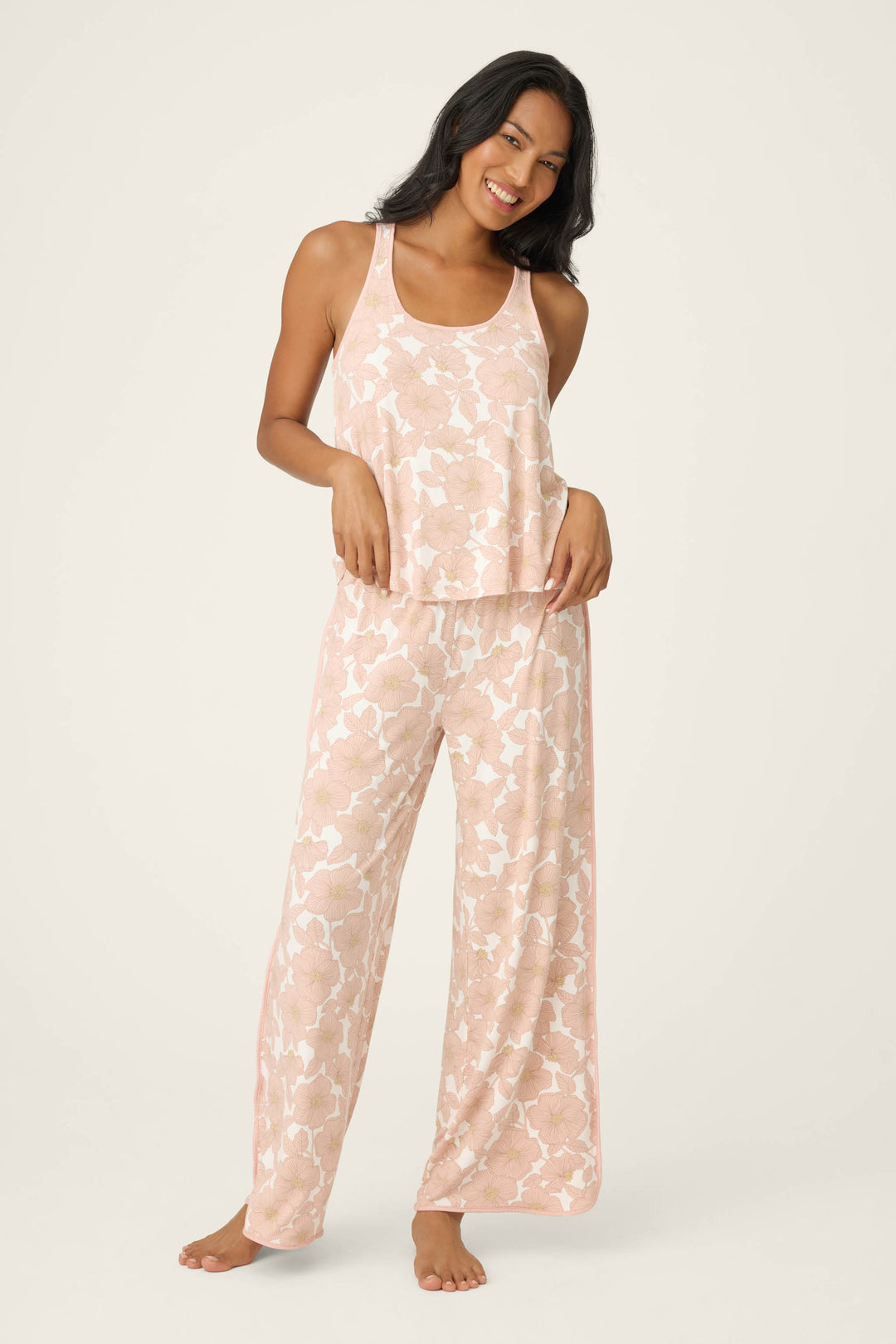 A woman stands barefoot against a plain background, smiling with one hand on her hip, wearing the P.J. Salvage Poppy Dreams Floral Tank Top and matching wide-leg pajama pants in pink and white.