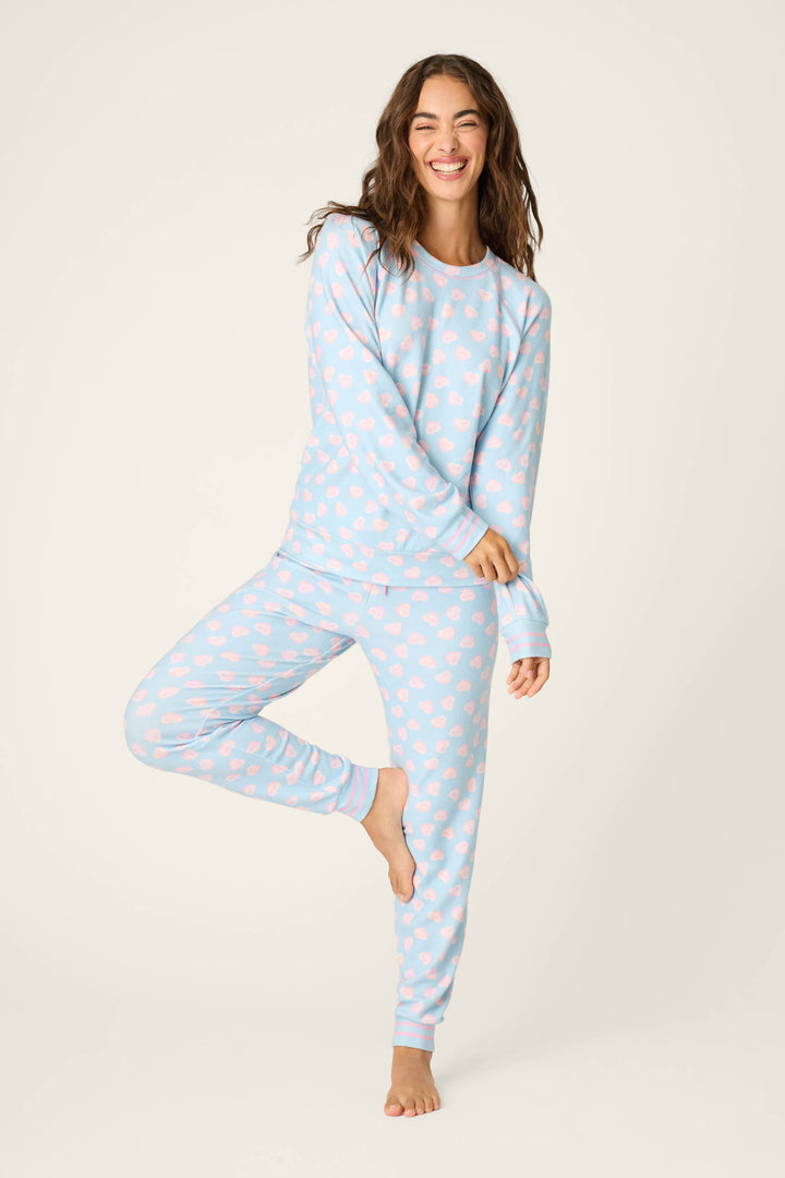 A woman stands on one leg, smiling, wearing the P.J. Salvage Sweetheart Love Hearts Print Peachy PJ Set against a plain light background.
