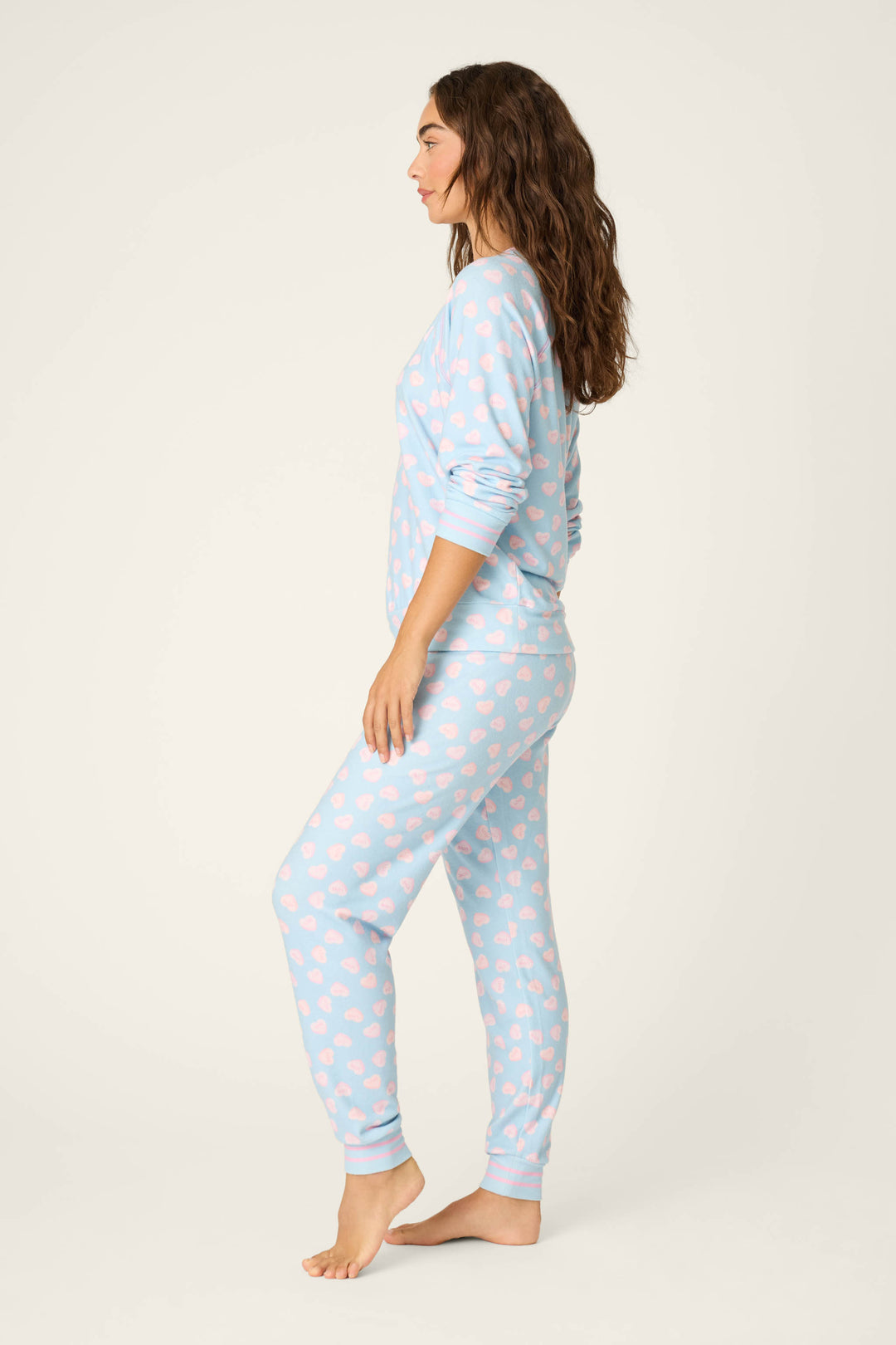 A woman stands barefoot in profile, wearing the P.J. Salvage Sweetheart Love Hearts Print Peachy PJ Set. The light blue long-sleeve pajamas feature a pink heart pattern, with her arms relaxed by her sides against a plain background.