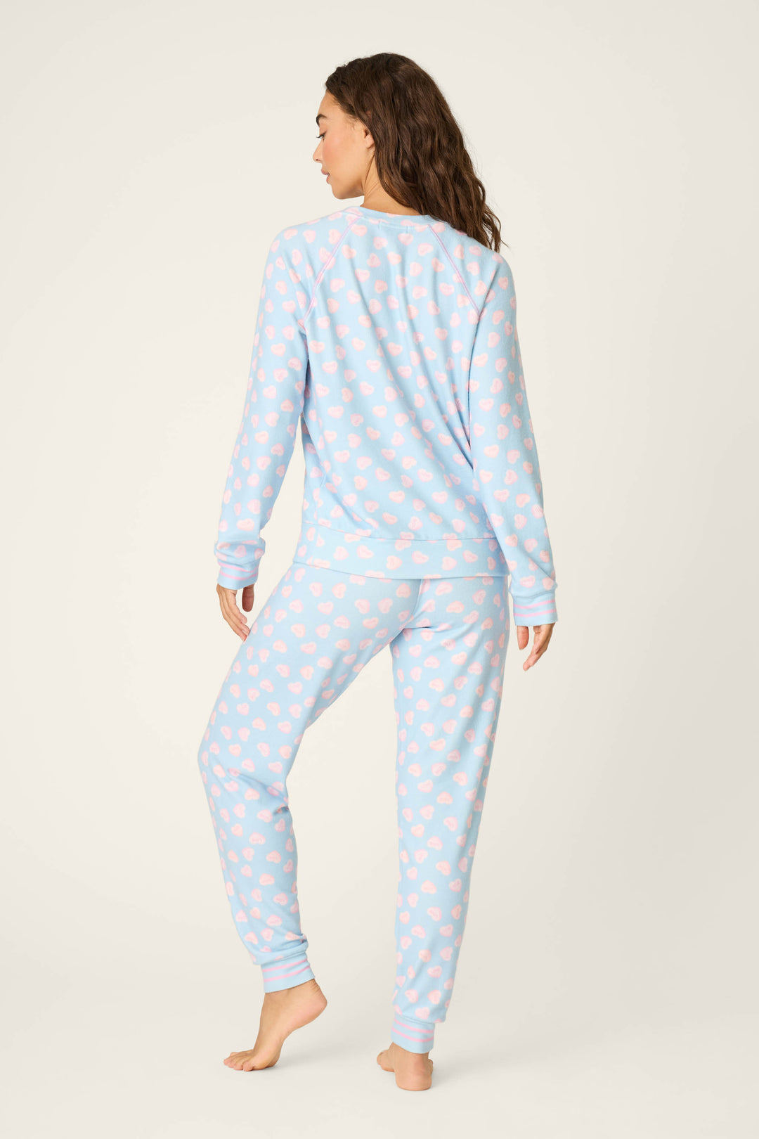 A person stands barefoot facing away, wearing the P.J. Salvage Sweetheart Love Hearts Print Peachy PJ Set—light blue with pink heart patterns and long sleeves and pants—set against a plain light background.