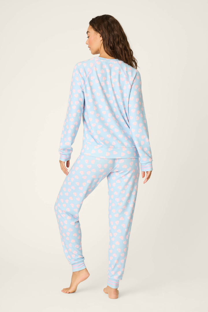 A person stands barefoot facing away, wearing the P.J. Salvage Sweetheart Love Hearts Print Peachy PJ Set—light blue with pink heart patterns and long sleeves and pants—set against a plain light background.