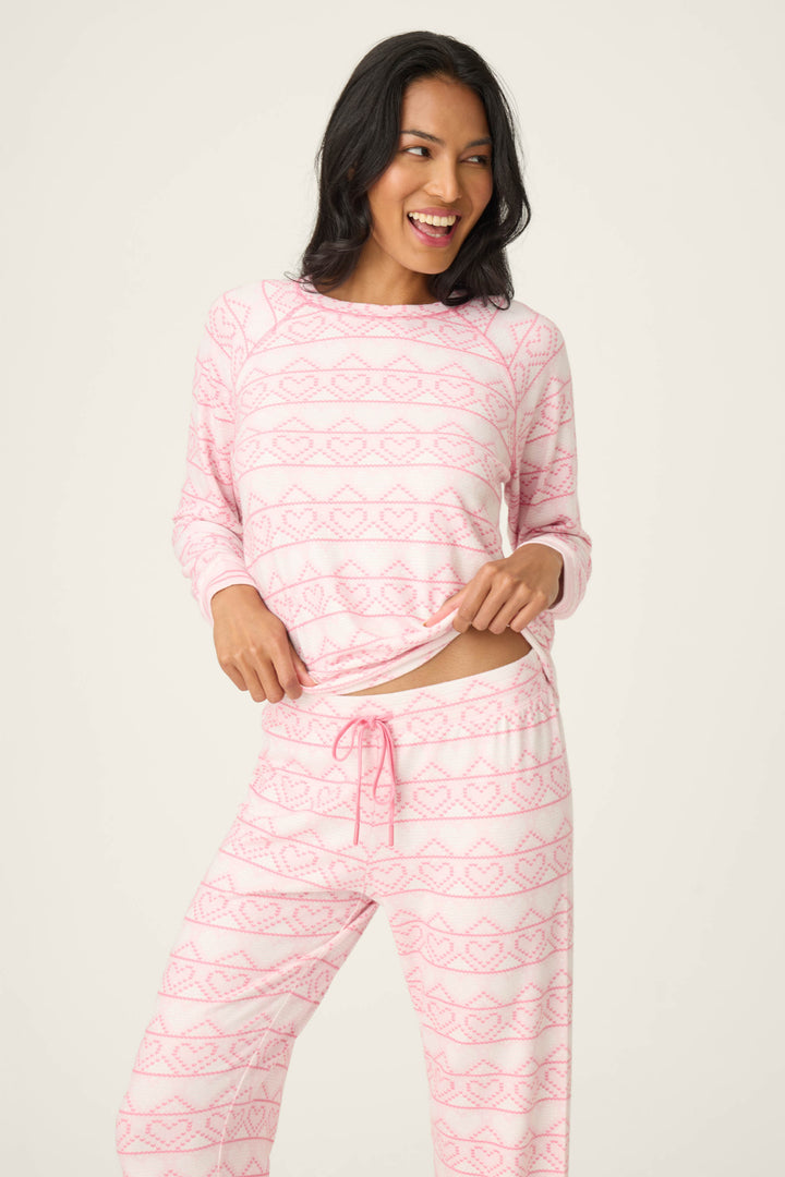 A woman smiles in the P.J. Salvage Fairisle Heart Print Peachy PJ Set, featuring a light pink color and subtle geometric heart pattern, holding up part of the top with one hand and posing against a plain light background.