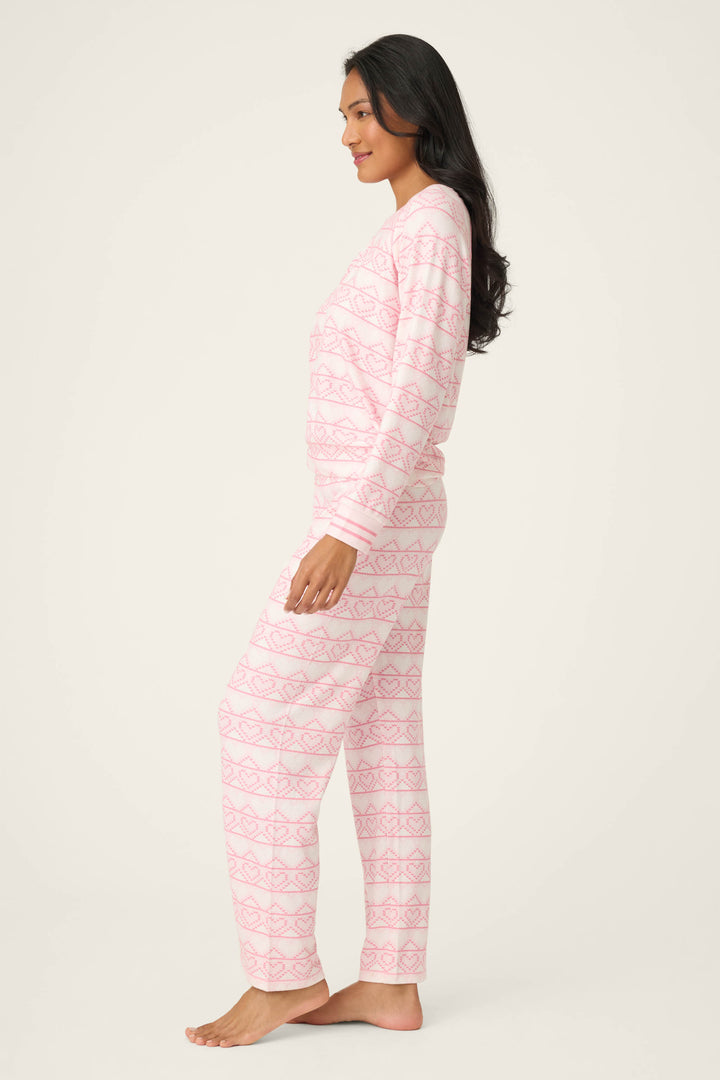 A woman stands barefoot in profile, wearing the P.J. Salvage Fairisle Heart Print Peachy PJ Set—a matching long-sleeve pink and white patterned pajama ensemble—against a plain light background.