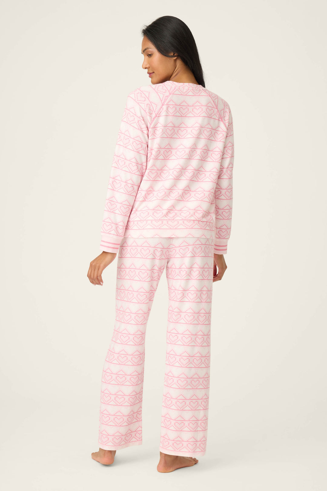A person stands barefoot facing away, wearing the P.J. Salvage Fairisle Heart Print Peachy PJ Set featuring a long-sleeve top and pants with a geometric pink and white pattern. The background is plain and light-colored.