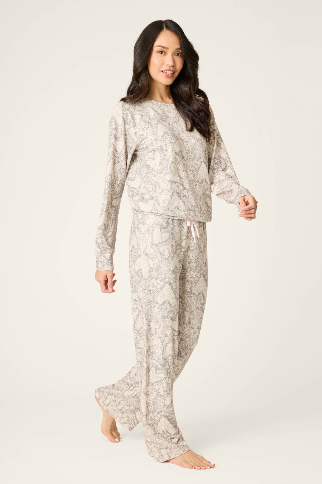 A person stands barefoot against a plain light background wearing P.J. Salvage's Snakeskin Heart Print Peachy PJ Set—light-colored, long-sleeve, and loose-fitting pajamas with a subtle heart and snakeskin pattern.