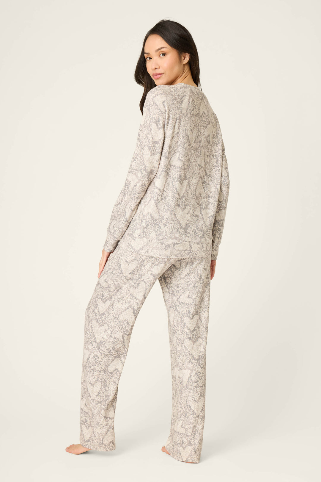 A person stands barefoot on a light surface, facing away in the P.J. Salvage Snakeskin Heart Print Peachy PJ Set—a matching long-sleeve top and pants featuring a light, textured snakeskin heart print pattern.