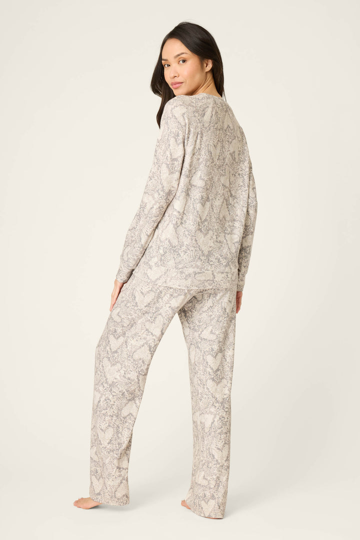 A person stands barefoot on a light surface, facing away in the P.J. Salvage Snakeskin Heart Print Peachy PJ Set—a matching long-sleeve top and pants featuring a light, textured snakeskin heart print pattern.