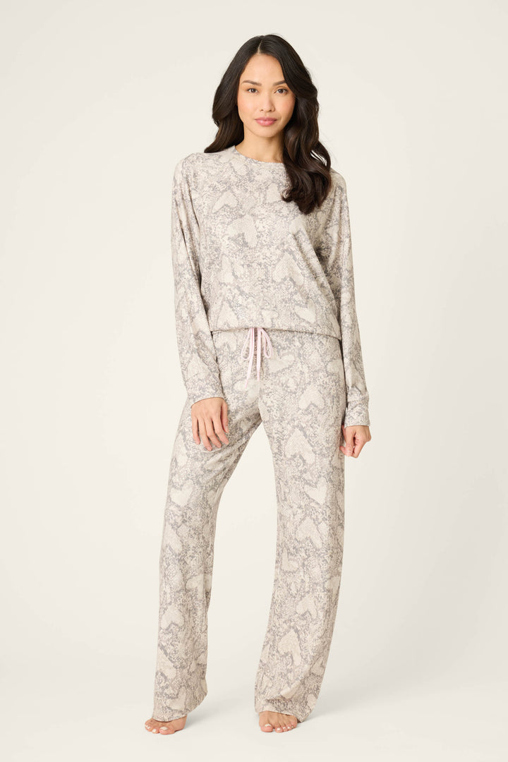A person stands facing forward, barefoot against a plain off-white background, wearing the P.J. Salvage Snakeskin Heart Print Peachy PJ Set—a matching long-sleeve top and pants featuring a light gray and white marbled pattern.