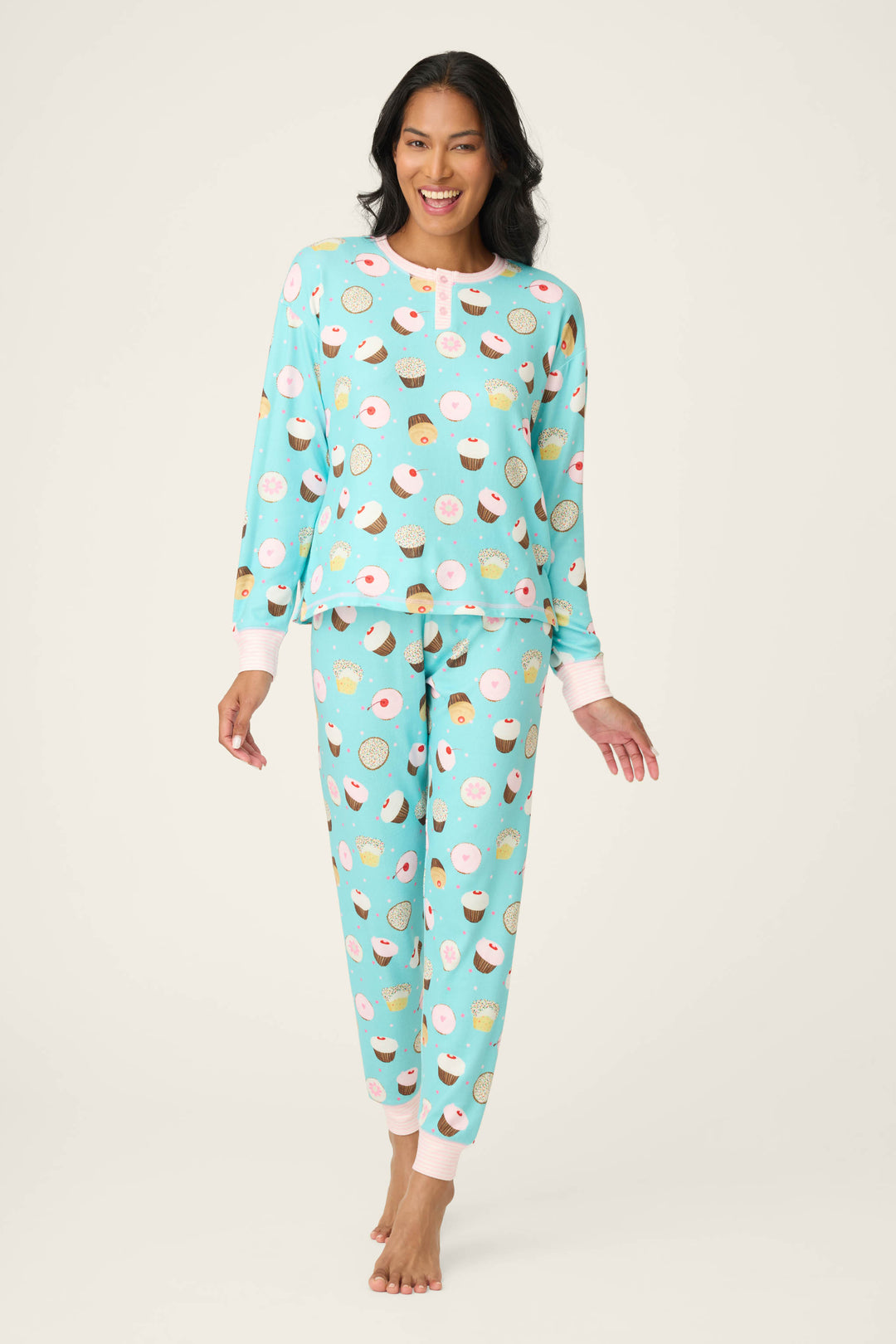A woman stands barefoot and smiling in the P.J. Salvage Sweet Life Cupcake Print PJ Set, featuring a long-sleeve top and full-length pants with a colorful cupcake pattern. The background is plain and light-colored.