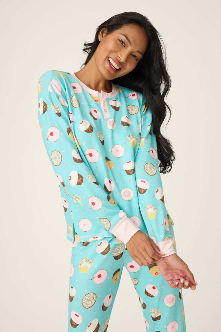 A woman smiles in the P.J. Salvage Sweet Life Cupcake Print PJ Set, featuring light blue pajamas with cupcake, donut, and sprinkle prints. She stands against a plain background with arms loosely crossed and head tilted.
