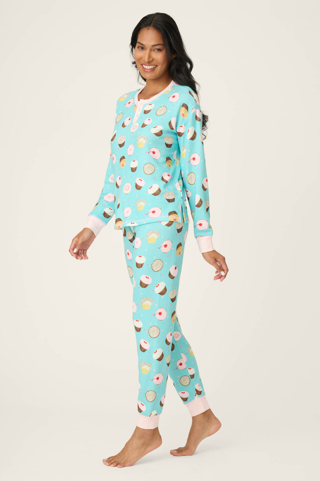 A smiling woman with long dark hair stands barefoot in a P.J. Salvage Sweet Life Cupcake Print PJ Set, featuring a colorful donut and cupcake pattern. She poses against a plain, light-colored background.